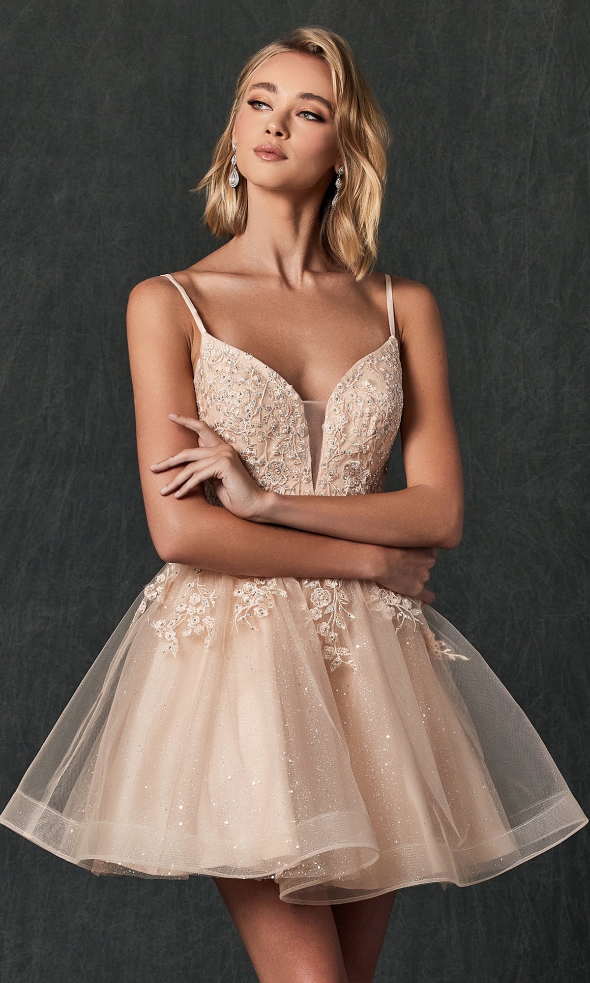 A woman models the Short Lace-Up Babydoll Homecoming Dress 859, a sleeveless beige party dress with intricate lace and sequin details, standing against a dark textured background.